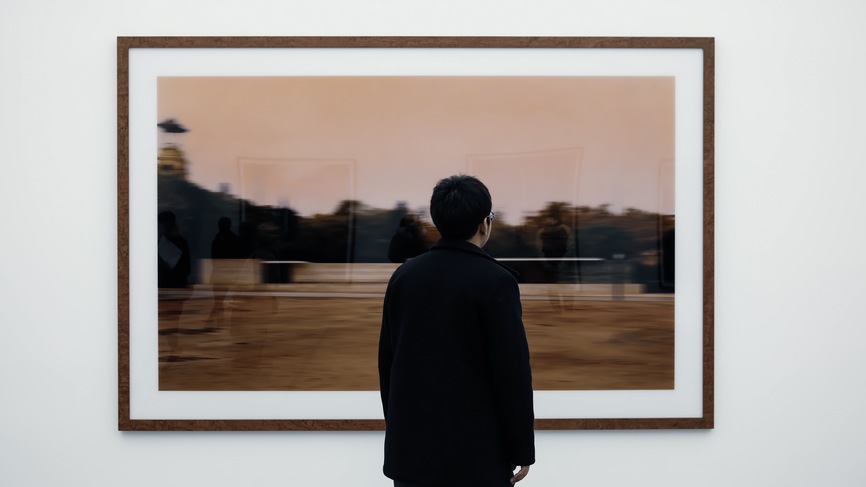 Shot from the back, a man is seen admiring a framed artwork at a gallery